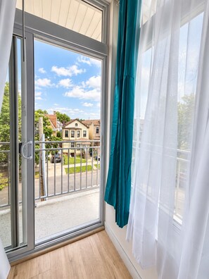 Une pièce dotée d’une porte-fenêtre coulissante donnant sur un balcon, d’un rideau blanc et offrant une vue sur des maisons et une rue.