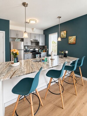 A modern kitchen with a marble island, teal chairs, and pendant lighting.