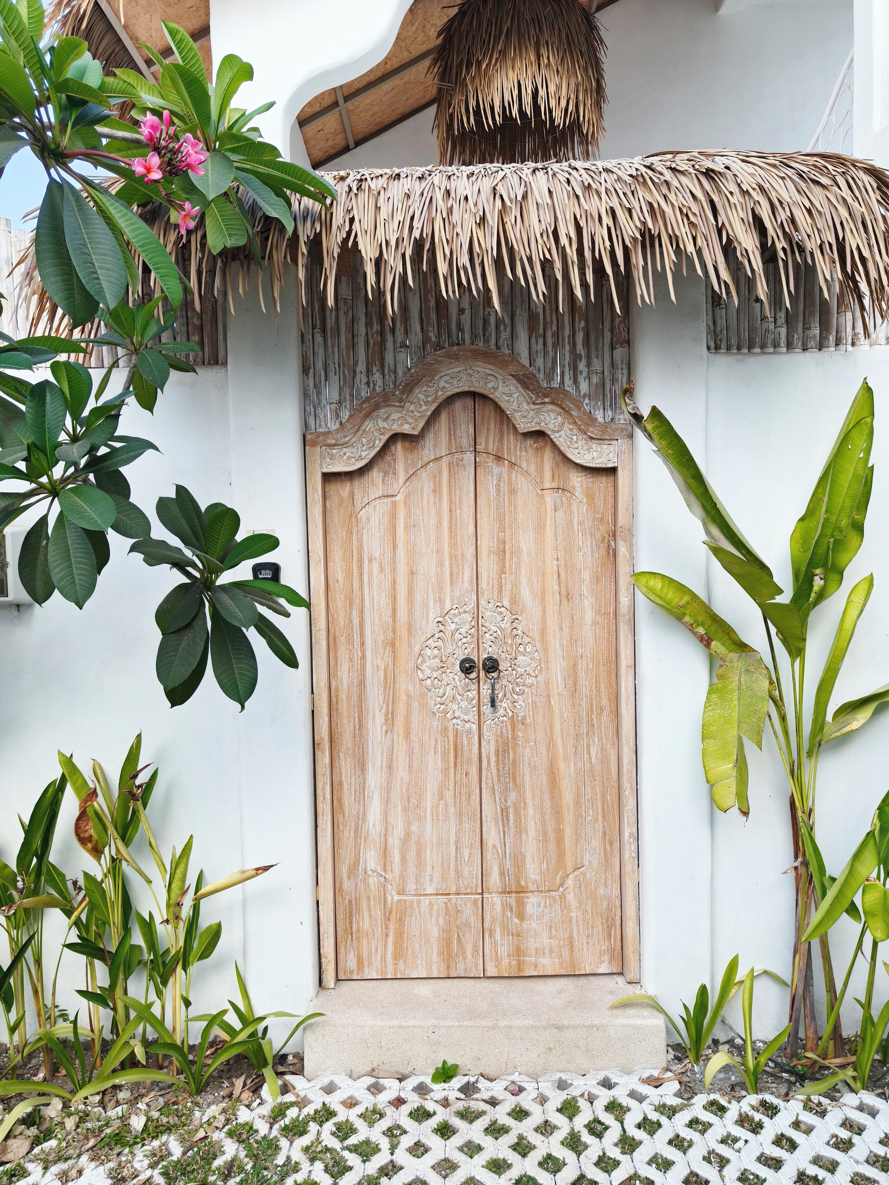 A wooden door with an ornate design, flanked by tropical plants and a thatched roof.
