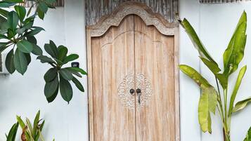 A wooden door with an ornate design, flanked by tropical plants and a thatched roof.