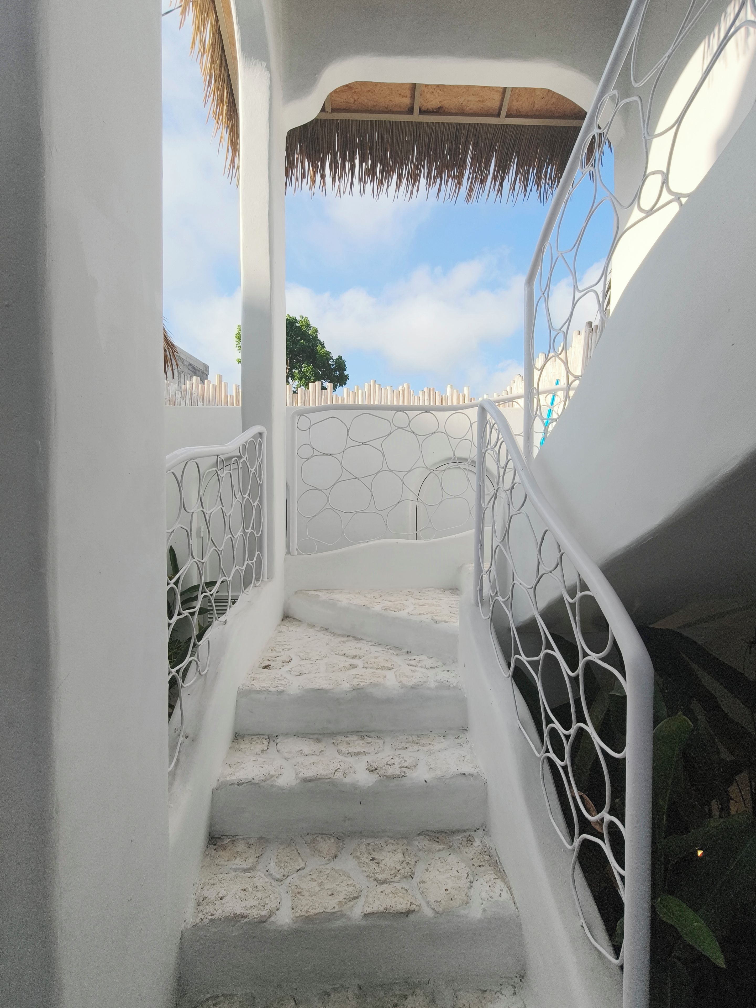 Una escalera con paredes blancas y techo de paja que lleva a un espacio al aire libre con vista al cielo y una cerca.