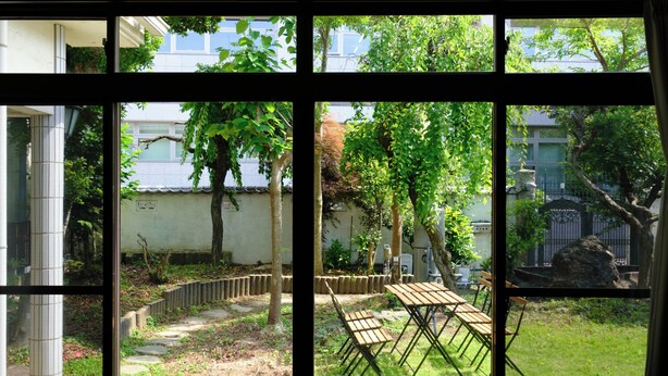 A view through a window of a courtyard with a wooden table and chairs, a stone path, and a fence. - Vrbo Property (hiratsuka yaezakityou)