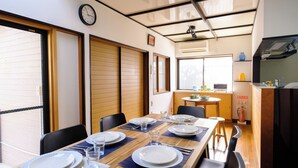 A dining area with a wooden table set for four, chairs, a clock on the wall, and a kitchen area in the background. - Vrbo Property (hiratsuka yaezakityou)