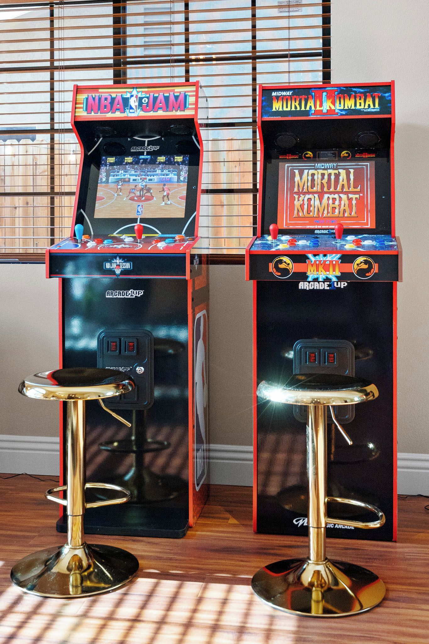 Two arcade machines, one for NBA JAM and the other for Mortal Kombat, placed on a wooden floor with a window in the background.