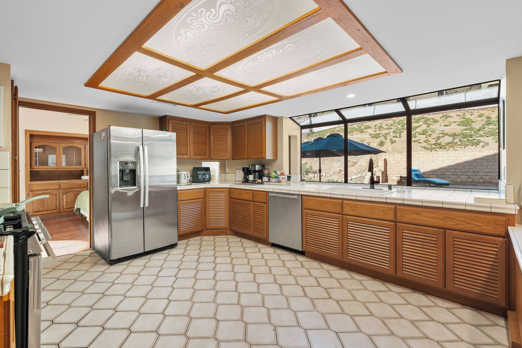 A modern kitchen with a stainless steel refrigerator, wooden cabinets, a tiled floor, and a view of a landscaped area through large windows.