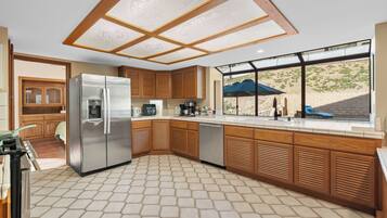 A modern kitchen with a stainless steel refrigerator, wooden cabinets, a tiled floor, and a view of a landscaped area through large windows.