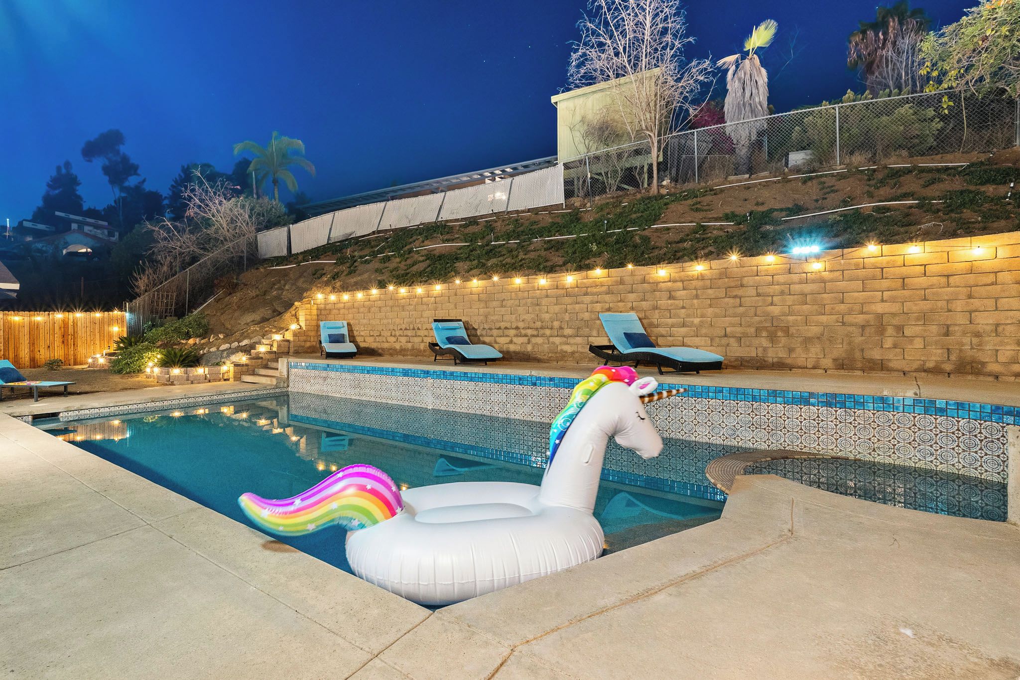 Una piscina con un flotador de unicornio, sillas de descanso y una pared de ladrillo con luces colgantes.