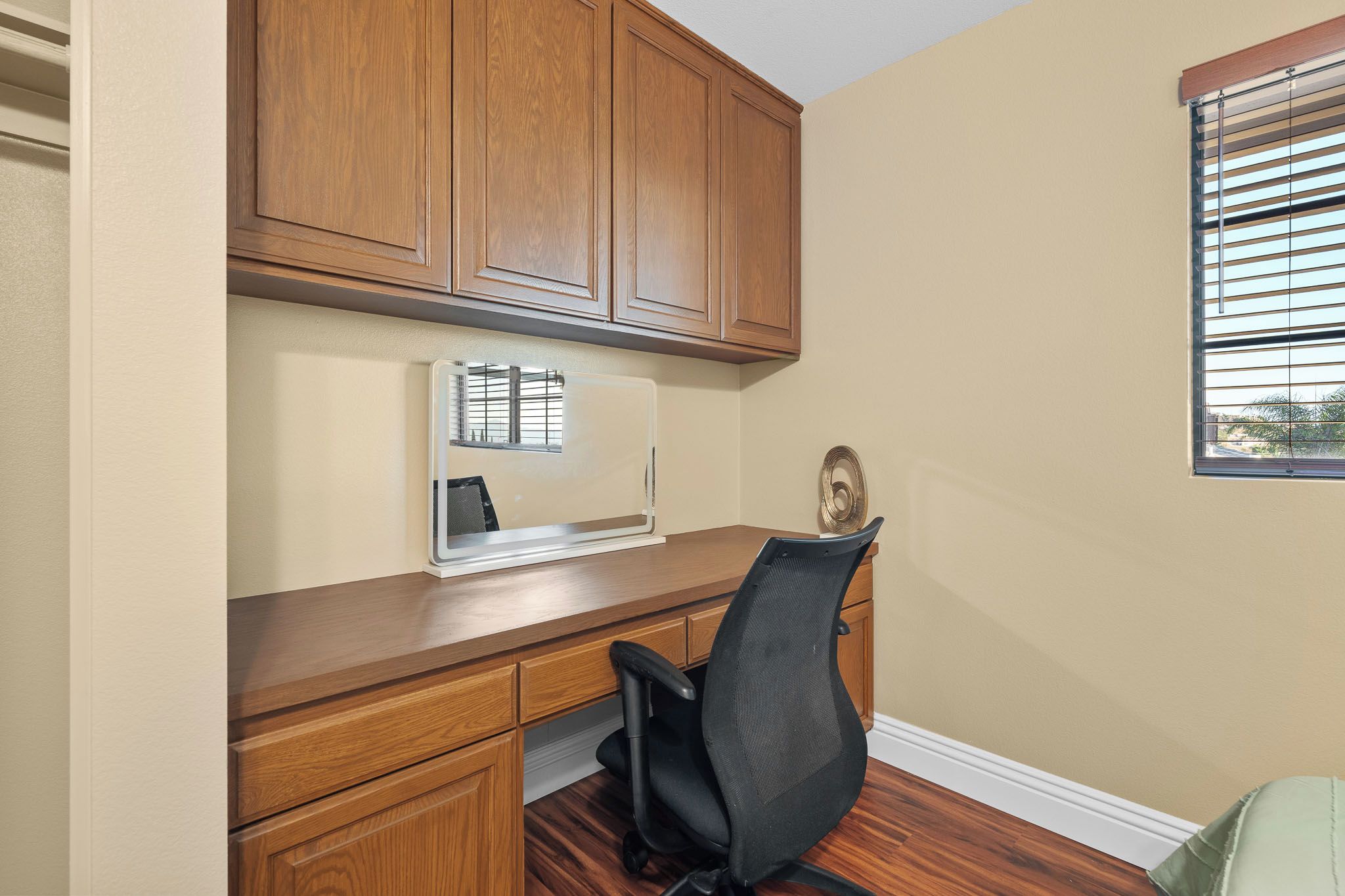 A home office with a wooden desk, a black office chair, and a window with blinds.