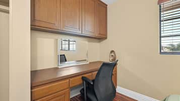 A home office with a wooden desk, a black office chair, and a window with blinds.