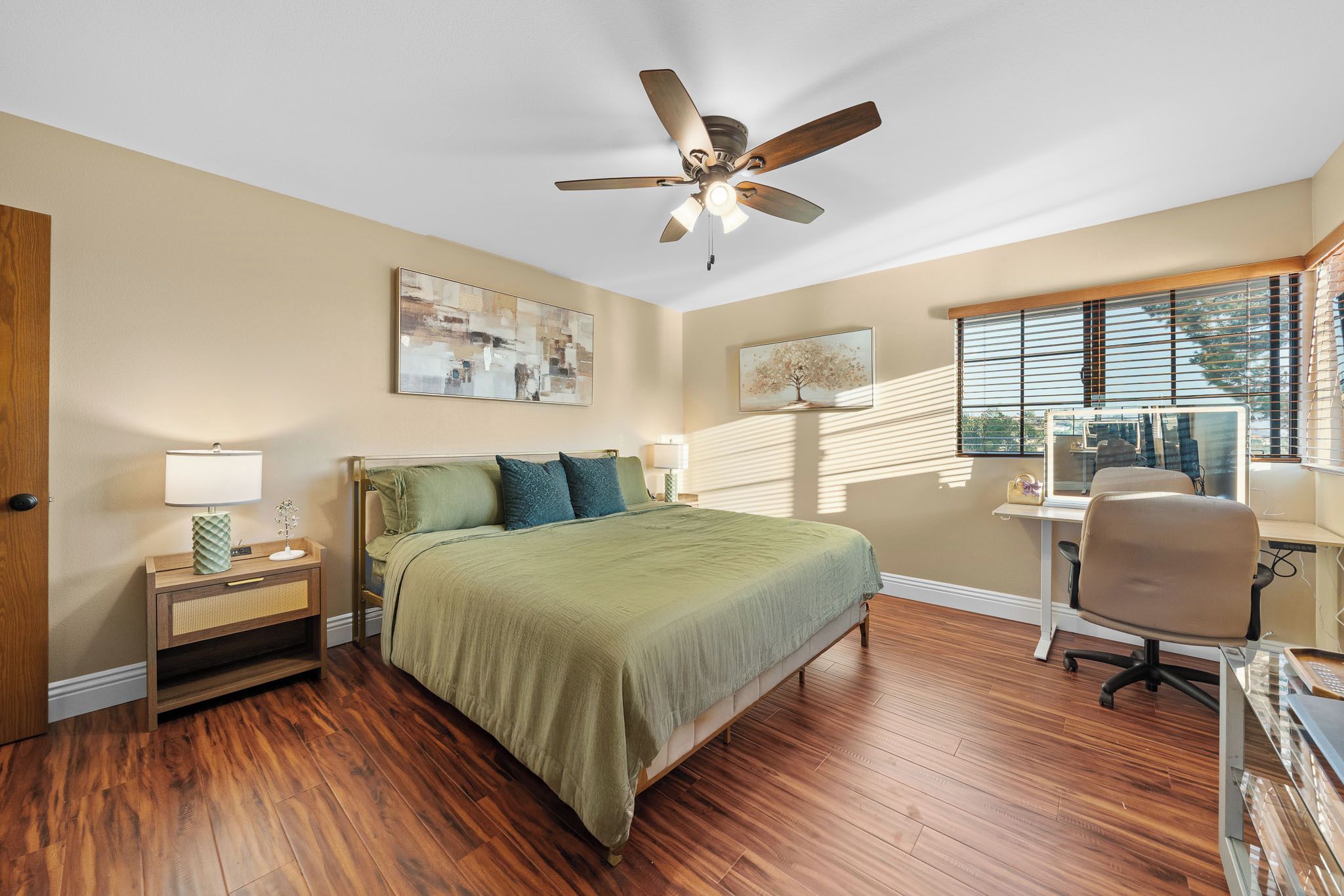A bedroom with a bed, bedside tables, a desk, a chair, a ceiling fan, and artwork on the walls.