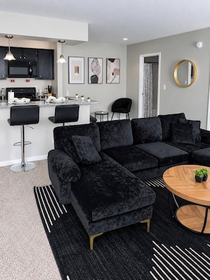 A modern living room with a sectional sofa, a round coffee table, and a kitchen area with a microwave and dining setup. - 3 mins from Arsenal/Bridge Street. (Huntsville)