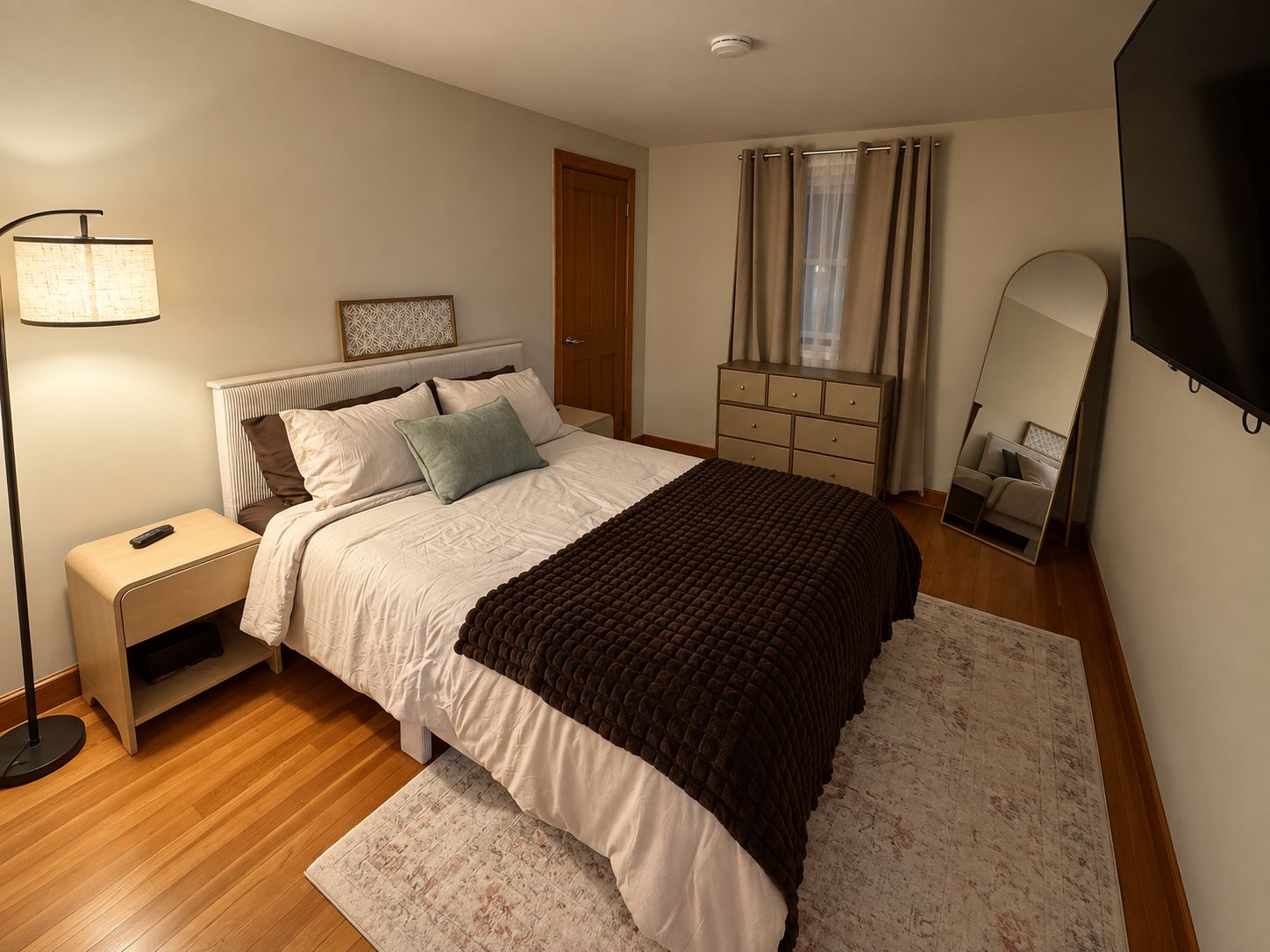 A bedroom with a bed, bedside table, lamp, dresser, mirror, and curtains.
