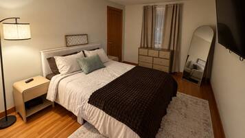 A bedroom with a bed, bedside table, lamp, dresser, mirror, and curtains.