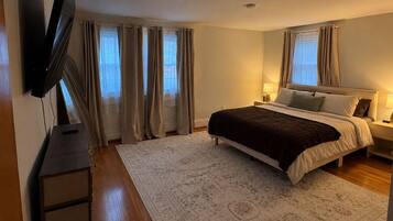 Ein Schlafzimmer mit einem Bett, einem flachbildfernseher an der Wand, einem Nachttisch mit Lampe und einem Fenster mit Vorhängen.