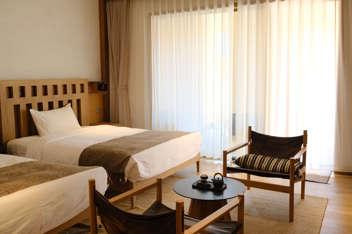 Habitación de hotel con dos camas, una silla, una mesita con tetera y ventana con cortinas.