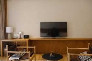 Sala de estar com televisão de tela plana na parede, móveis de madeira, uma mesinha com um objeto decorativo e uma luminária em um criado-mudo.