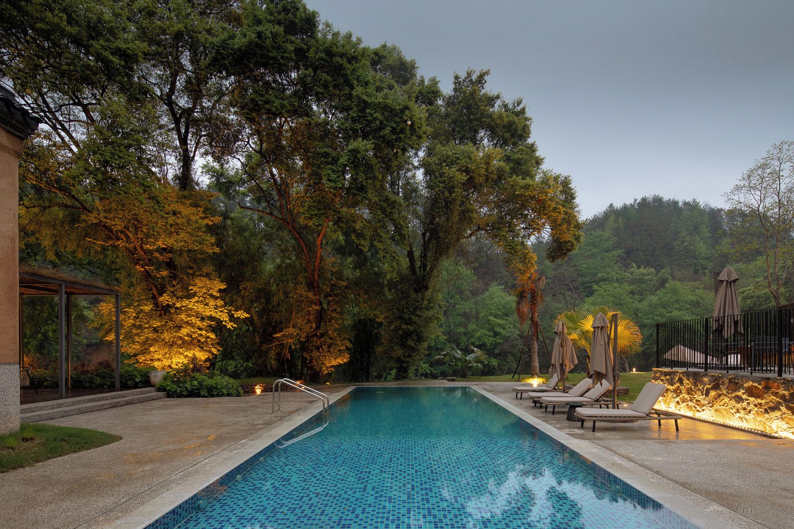 Una piscina con sillas de descanso y una pérgola, rodeada de árboles y un muro de piedra.
