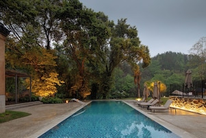 A swimming pool with lounge chairs and a pergola, surrounded by trees and a stone wall.