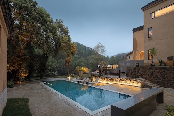 A swimming pool with lounge chairs, surrounded by trees and a stone wall.