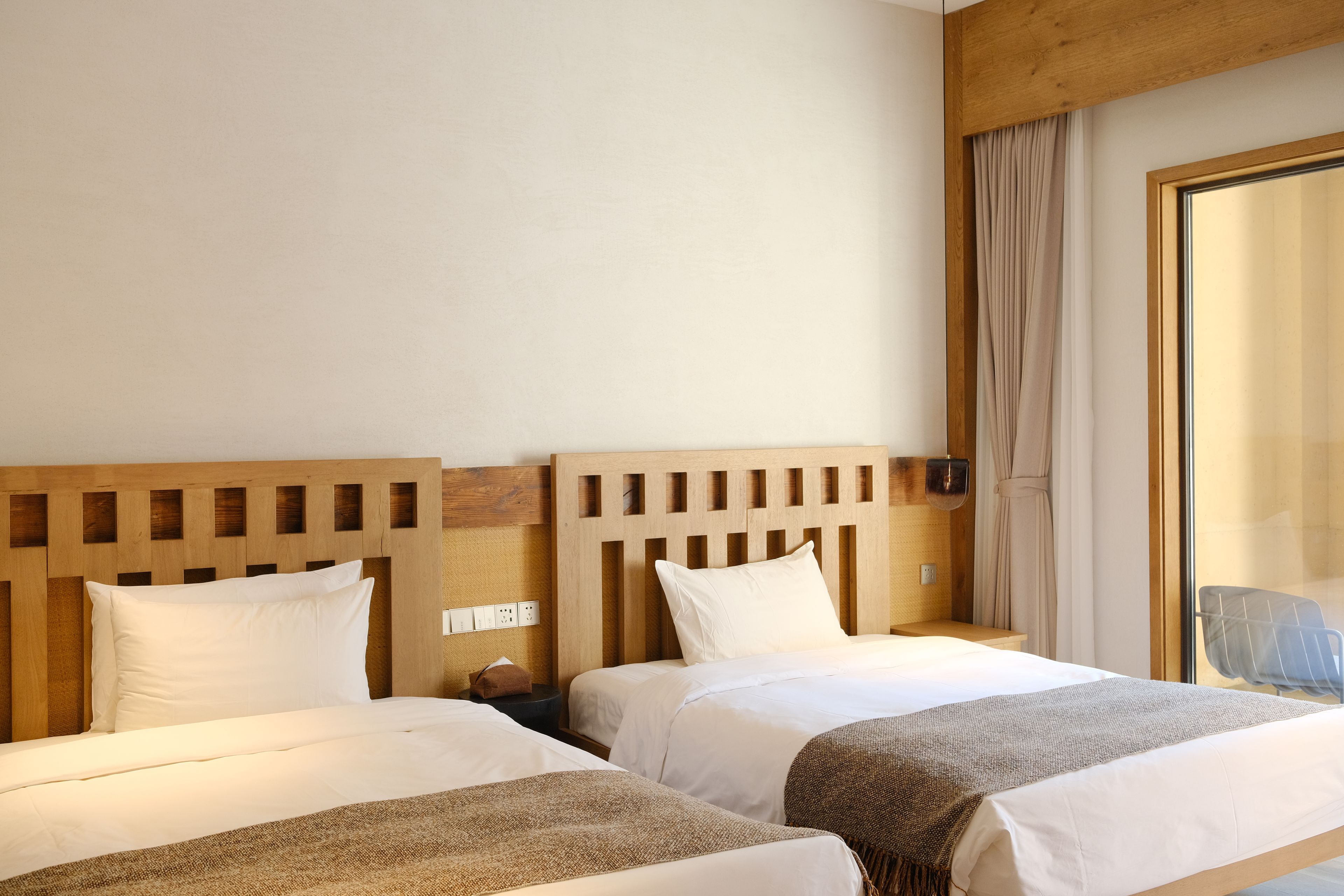 A hotel room with two beds, wooden headboards, a window with curtains, and a view of the outside.