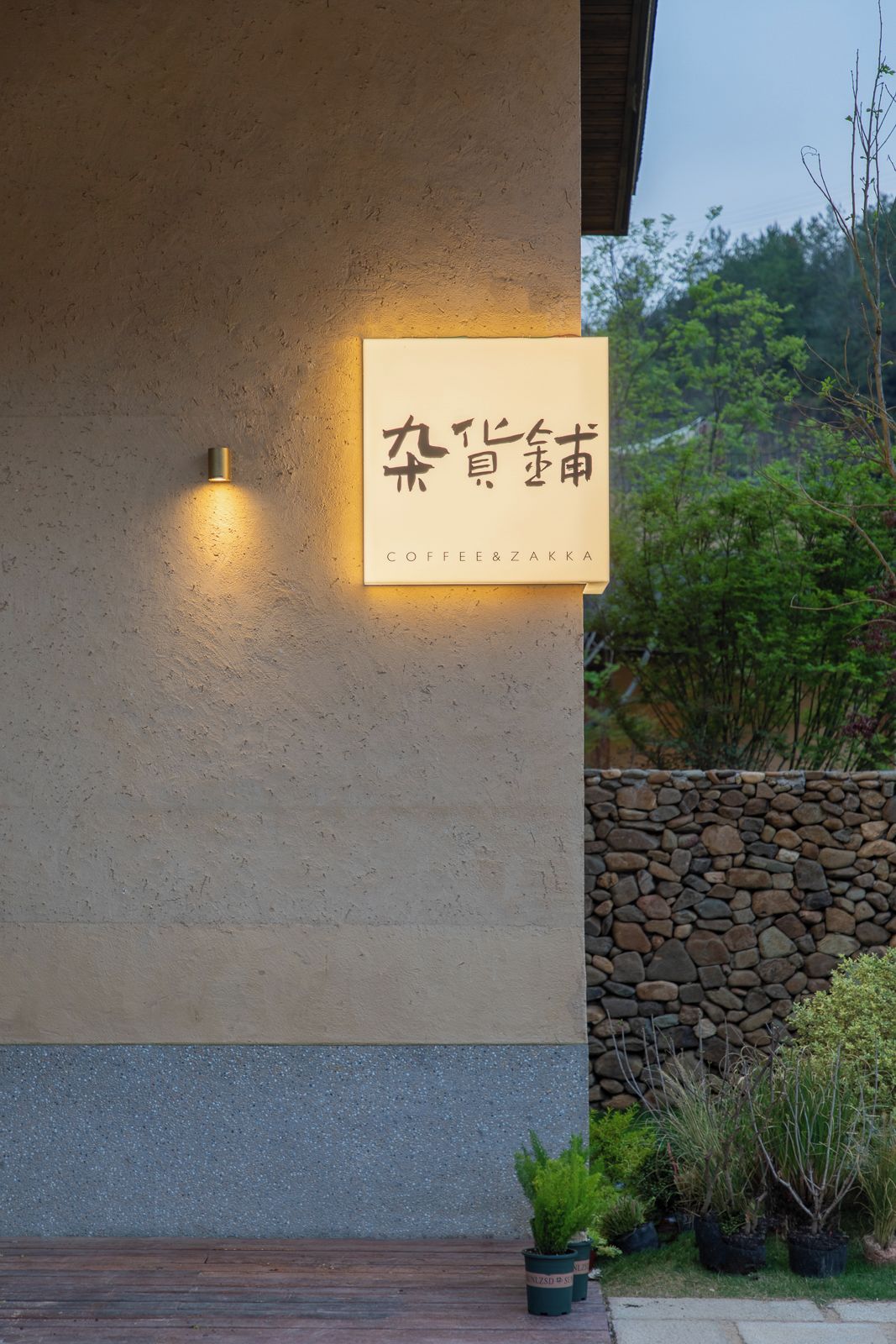 Un letrero con caracteres chinos y 'Coffee & Zakka' escrito debajo, sujeto a una pared de piedra con plantas de fondo.
