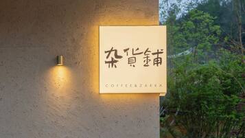 Un letrero con caracteres chinos y 'Coffee & Zakka' escrito debajo, sujeto a una pared de piedra con plantas de fondo.