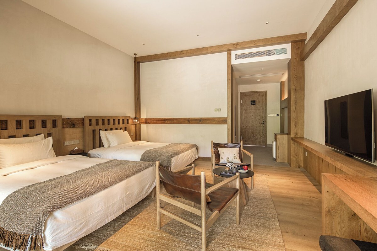 Habitación de hotel con dos camas, cabecera de madera, televisor y una pequeña mesa con una silla.