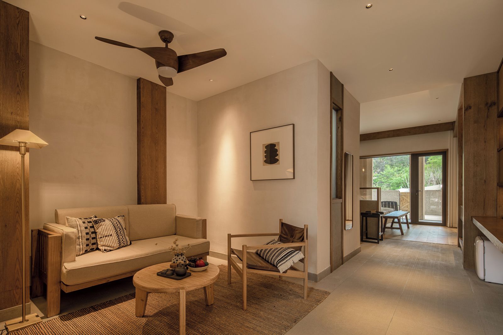 A modern living room with a sofa, a coffee table, and a chair. There is a ceiling fan, a framed picture on the wall, and a view of the outdoors through a glass door.