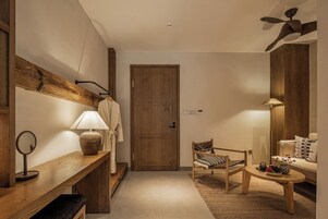 Habitación de hotel con puerta y banco de madera, una silla, una mesita, una lámpara, un espejo y un sofá.