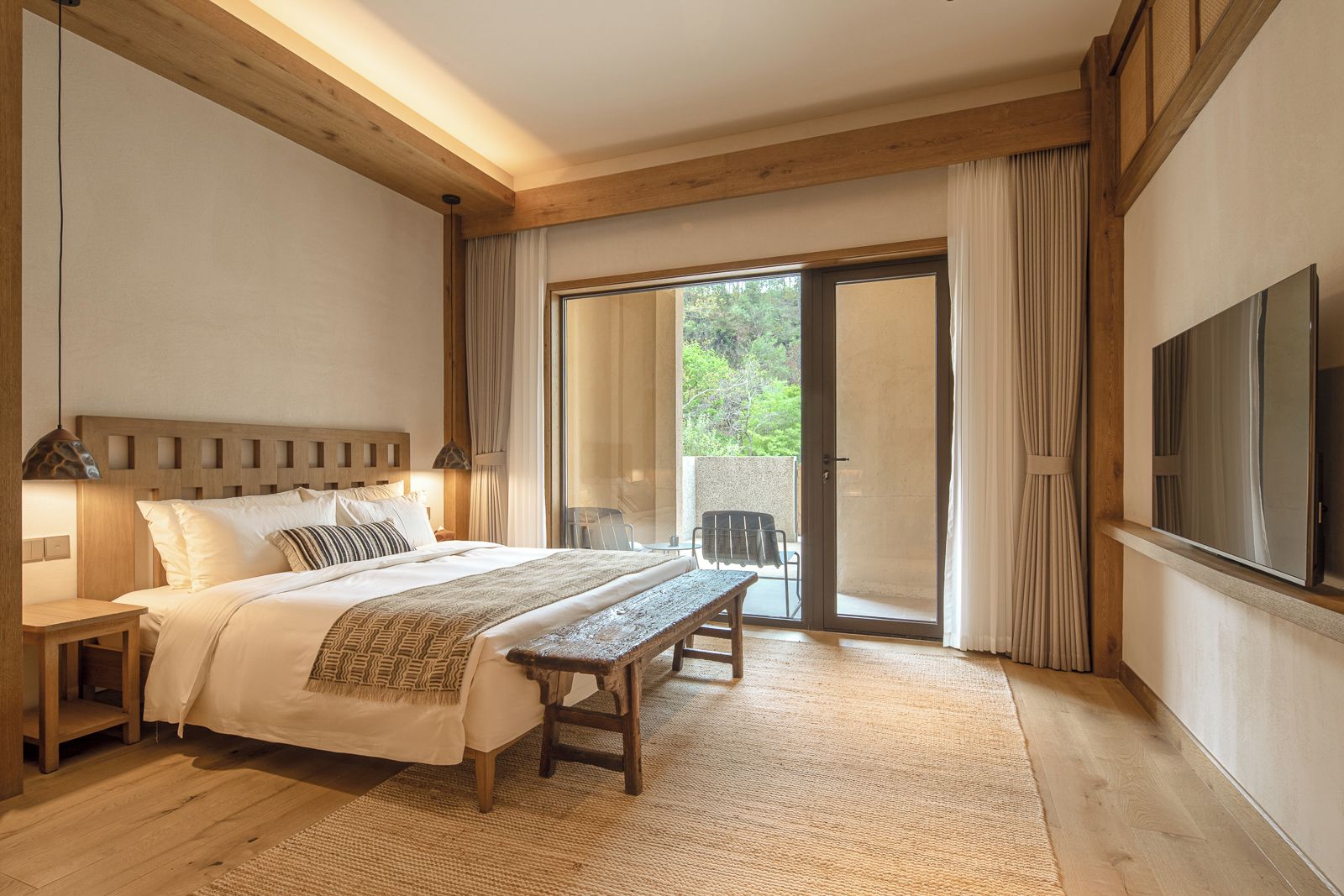 A bedroom with a bed, bedside table, bench, and a TV mounted on the wall.