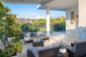 A modern outdoor seating area with wicker furniture, a glass balustrade, and lush greenery. - Casa Brio (Badung)