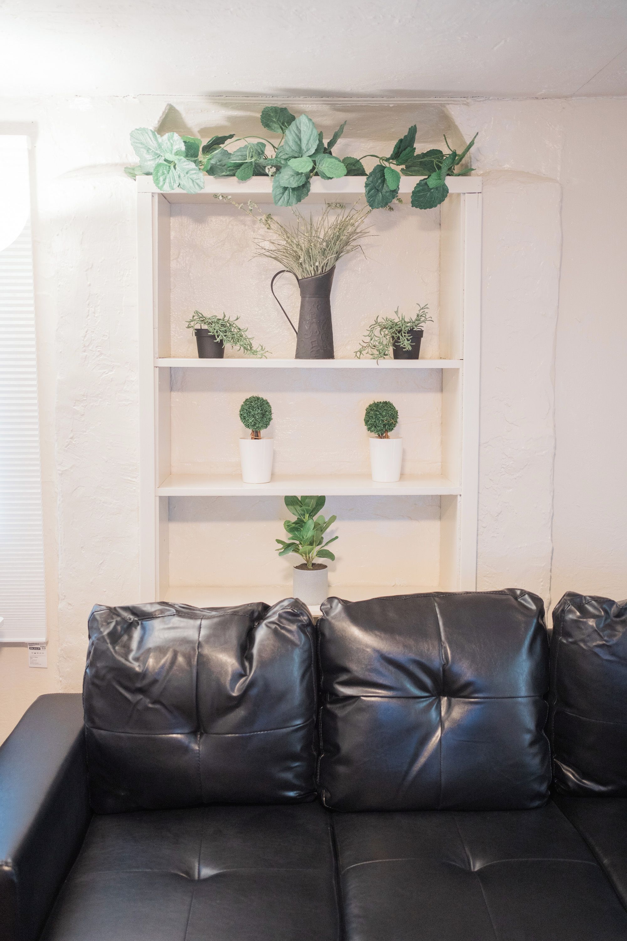A black leather sofa with three cushions, a white shelving unit with potted plants, and a white wall.