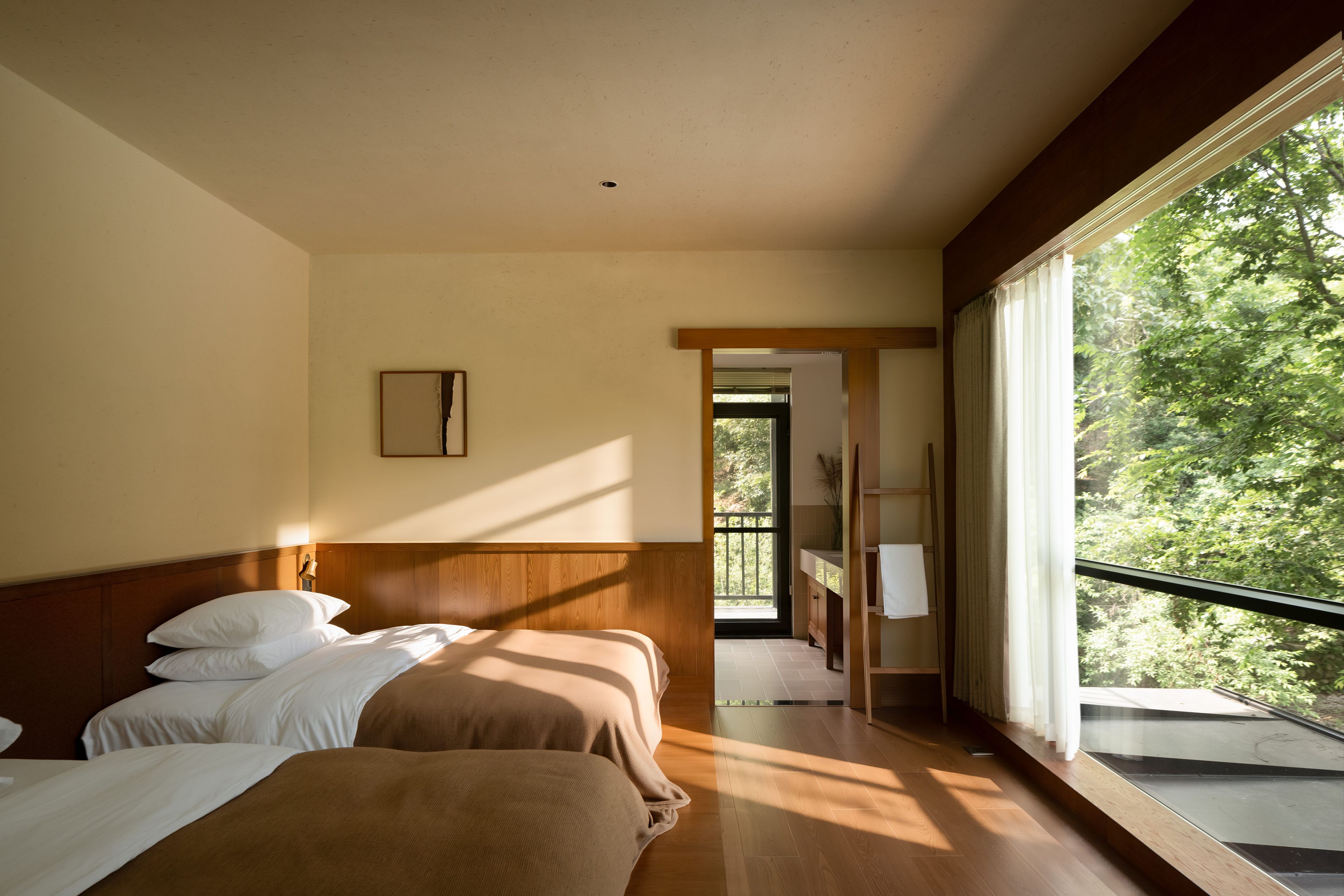 A hotel room with a large window, a bed, a bedside table, and a view of greenery outside.