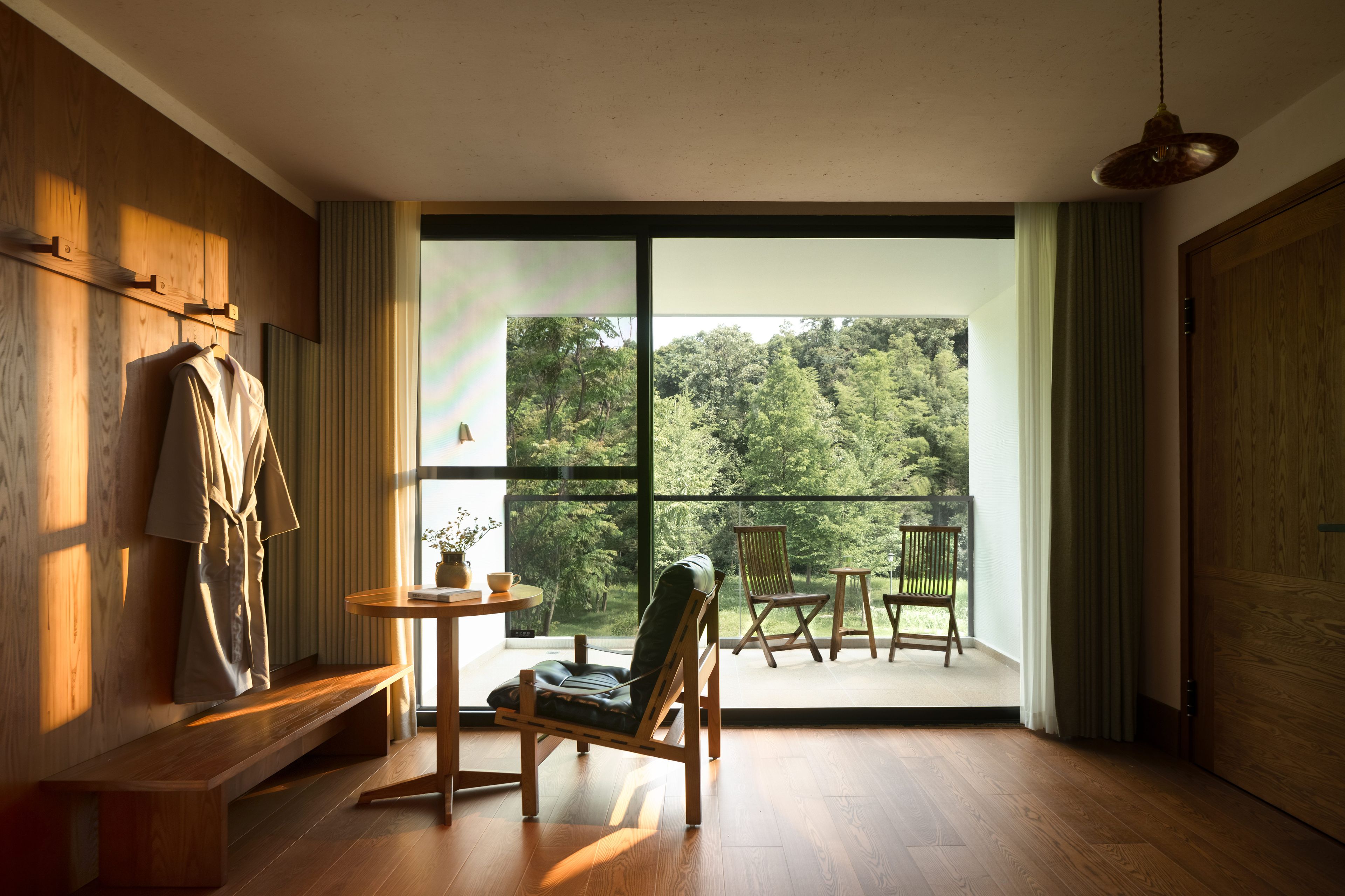 Una habitación con piso de madera, un banco, una mesa con un florero y una taza, una silla y vistas a un bosque a través de amplios ventanales.