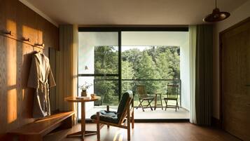 Una habitación con piso de madera, un banco, una mesa con un florero y una taza, una silla y vistas a un bosque a través de amplios ventanales.