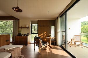 A modern interior with a bed, a wooden desk, a chair, and a large window offering a view of greenery.