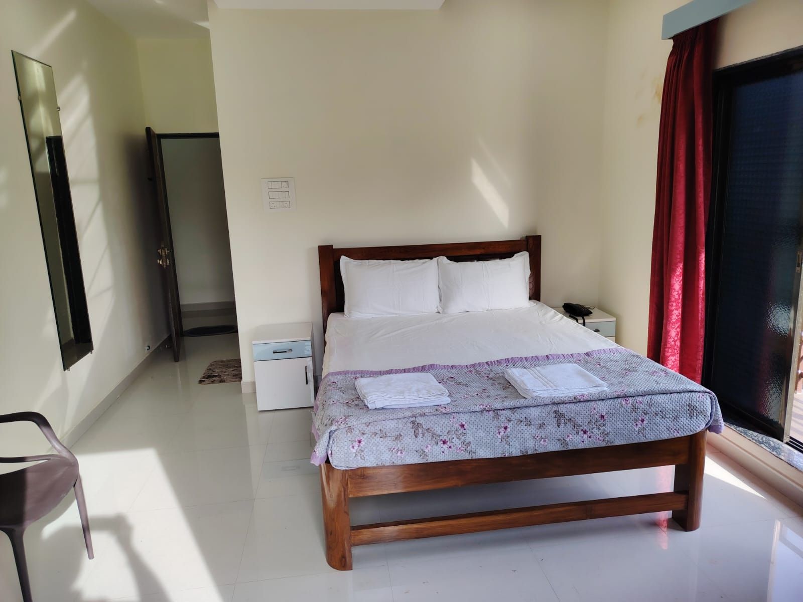 A neatly made bed with a floral-patterned bedspread and two folded towels, a bedside table with a lamp, a small white nightstand, and a mirror on the wall.