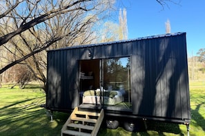 A small, dark-colored mobile home with a sliding door, a bed inside, and a wooden ramp leading up to it.