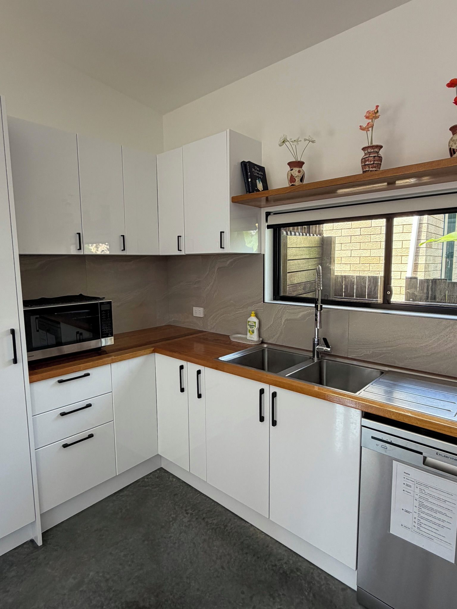 A modern kitchen with white cabinets, a wooden countertop, a built-in dishwasher, and a window with a view of a brick wall.