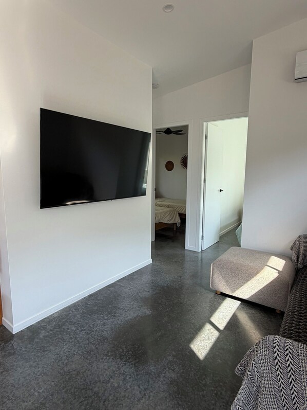 Una sala moderna con un televisor de pantalla plana montado en la pared, una alfombra gris, un sofá y una cama en la habitación contigua.