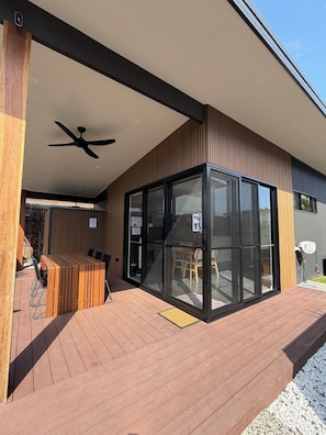 Un espacio exterior moderno con terraza de madera, ventilador de techo y zona de estar con paredes de vidrio.
