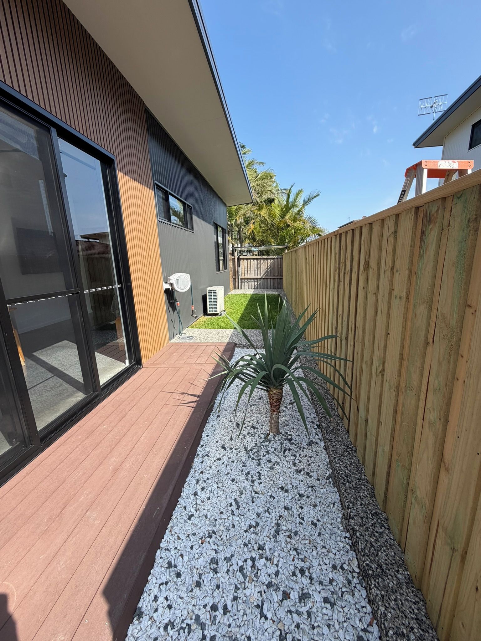 A modern house with a wooden deck, a fenced garden area, and a palm plant.