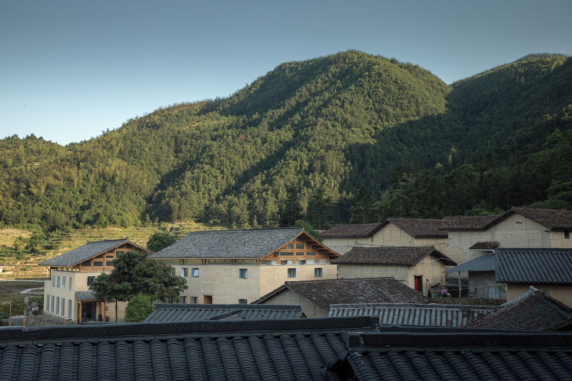A traditional village with buildings featuring sloped roofs, set against a backdrop of a forested mountain.