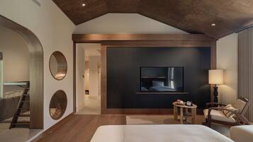 A modern hotel room with a large bed, a flat-screen TV mounted on the wall, a small table with a lamp, and a chair.