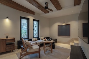 Uma sala de estar moderna com um sofá, cadeiras, uma mesa de centro e uma lareira. Há duas janelas com molduras de pedra e um ventilador de teto.
