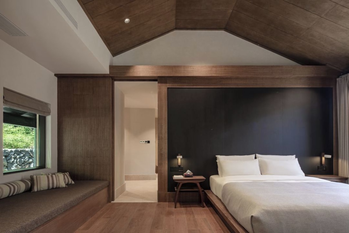 A modern bedroom with a large bed, a seating area with cushions, and a view of greenery through a window.
