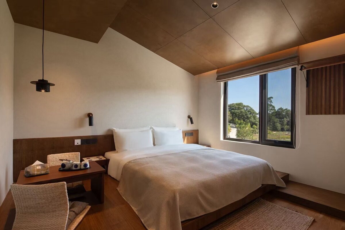 A modern hotel room with a large bed, a desk, a chair, and a window with a view of trees.
