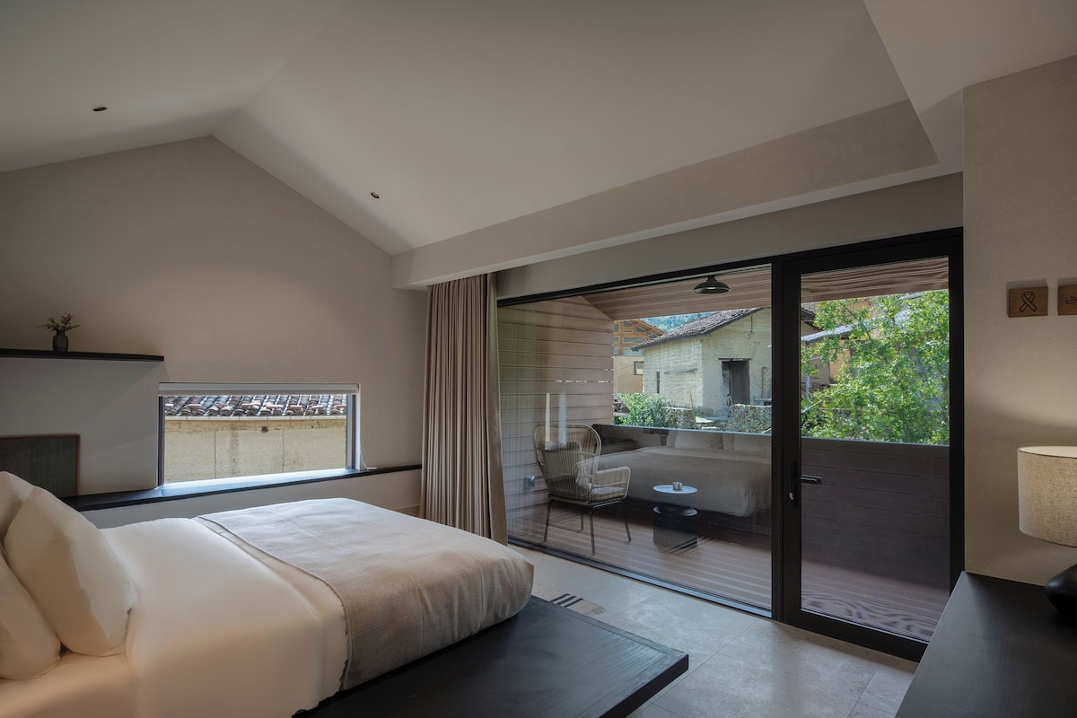 A modern hotel room with a bed, a small table, a chair, and a view of the outside through a sliding glass door.