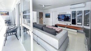 A modern living room with a grey sofa, a flat-screen TV, and a balcony with a table and chairs. - B8 - 2 Bedrooms Serviced Apartment with pool view, Karon Beach (Tambon Karon)