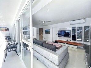 A modern living room with a grey sofa, a flat-screen TV, and a balcony with a table and chairs. - B8 - 2 Bedrooms Serviced Apartment with pool view, Karon Beach (Tambon Karon)